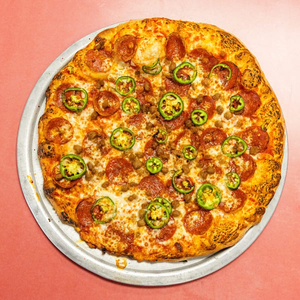 Whole pepperoni, sausage, and jalapeño pizza on a metal tray against a pink background.