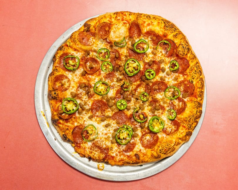 Whole pepperoni, sausage, and jalapeño pizza on a metal tray against a pink background.