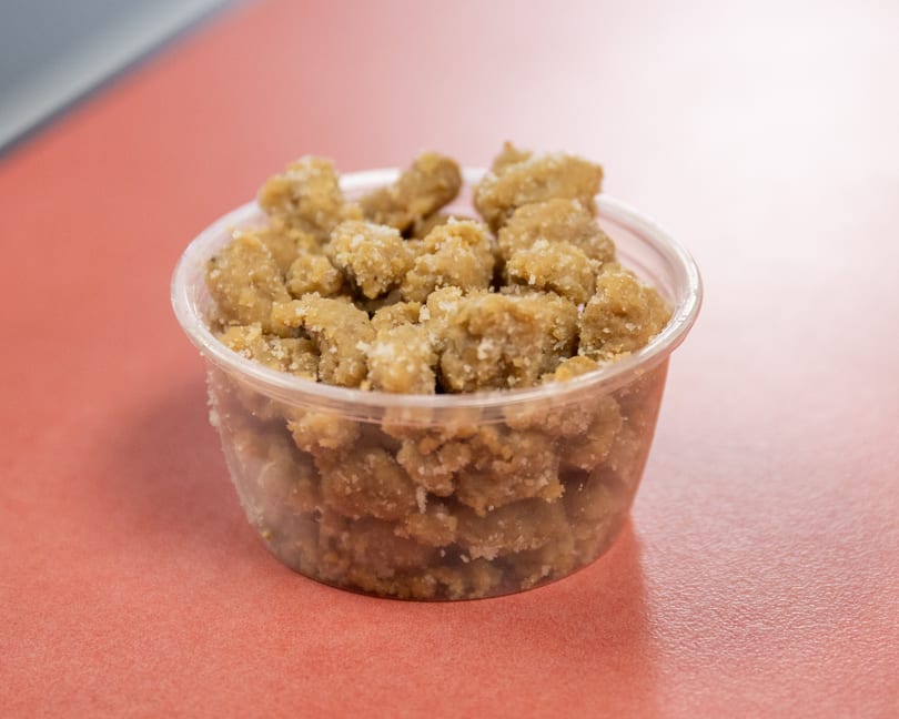Sugary brown dessert crumbles in a clear plastic container on a reddish-orange table.