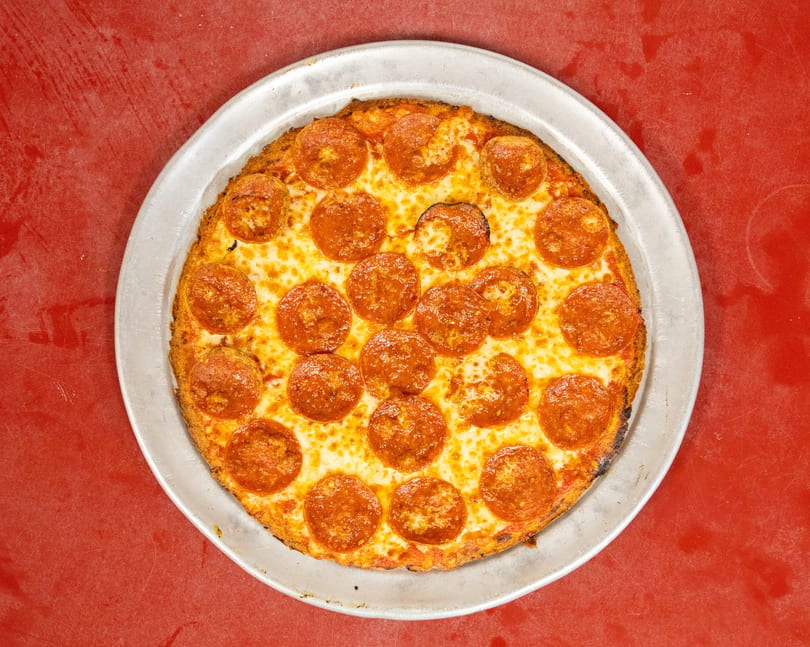 Whole pepperoni pizza with melted cheese on a metal pan, against a red background.