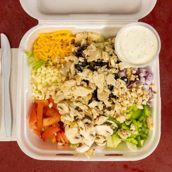 Takeout salad with chicken, shredded cheese, various vegetables, dressing, and plastic cutlery.