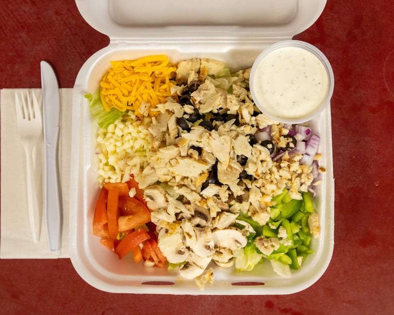 Takeout salad with chicken, shredded cheese, various vegetables, dressing, and plastic cutlery.