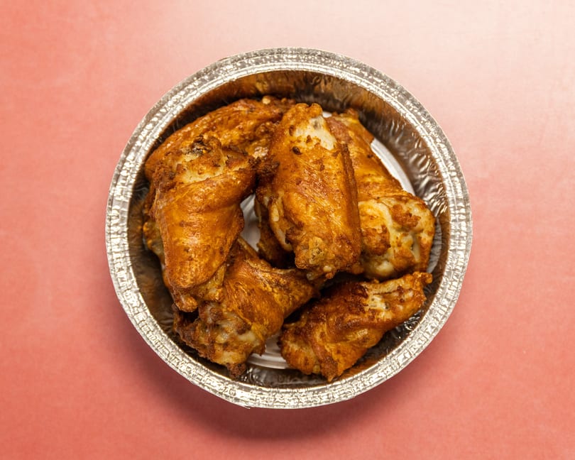 Golden-brown chicken wings in a round foil container on a pink background, viewed from above.