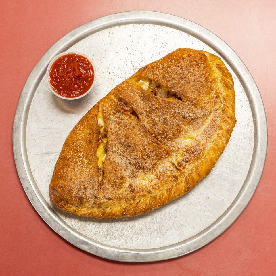 Golden brown calzone with cheese sprinkles and a side of marinara sauce on a metal pan.