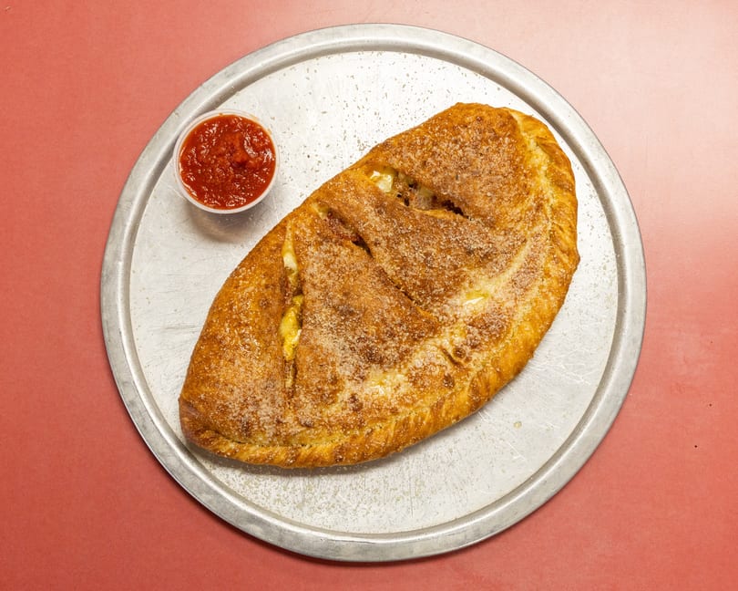 Golden brown calzone with cheese sprinkles and a side of marinara sauce on a metal pan.