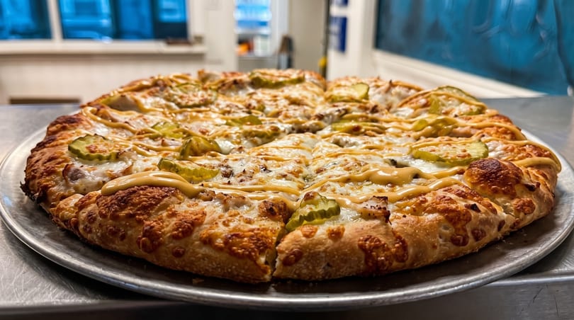 A whole pizza with pickles and a mustard drizzle, sliced and ready to eat.